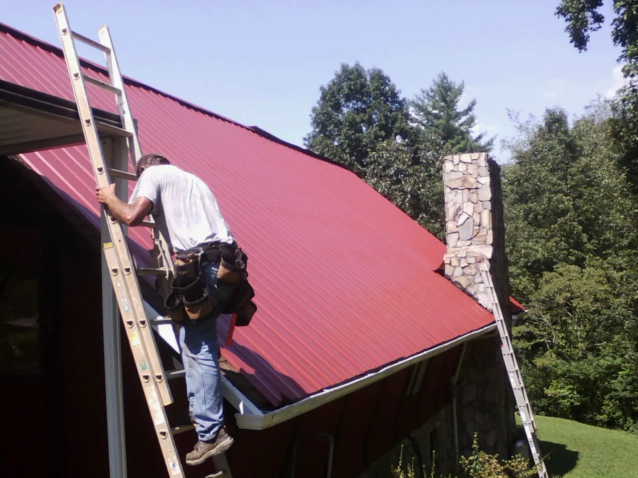 Roofer on a ladder beside a red metal roof and stone chimney — Soffit & Fascia Repair by Roofing Brothers in Pinon Hills